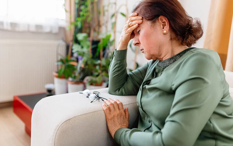 Eine ältere Frau sitzt auf einem Sofa, fasst mit der linken Hand an die rechte Seitenlehne, während sie mit der rechten Hand ihren Kopf abstützt.