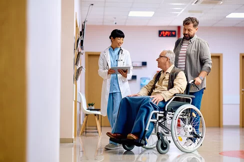 Ein älterer Mann im Rollstuhl wird von einem jüngeren Mann unterstützt, während eine Ärztin mit Unterlagen in ihren Händen daneben steht.