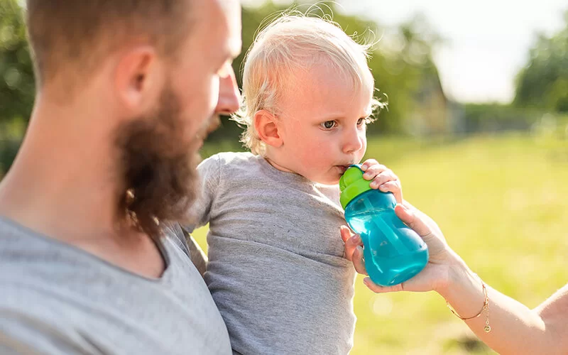 Ein Vater hält einen Jungen auf dem Arm, der eine Nuckelflasche mit Wasser gereicht bekommt.