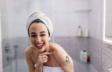 Eine Frau mit einem weißen Handtuch um den Körper und einem weiteren Handtuch um den Kopf steht in einem Badezimmer. Die Person hält eine Hand an das Kinn und hat ein sichtbares Tattoo auf der Schulter. Im Hintergrund sind Fliesen, ein beleuchteter Spiegel und mehrere Flaschen auf Glasablagen zu sehen.