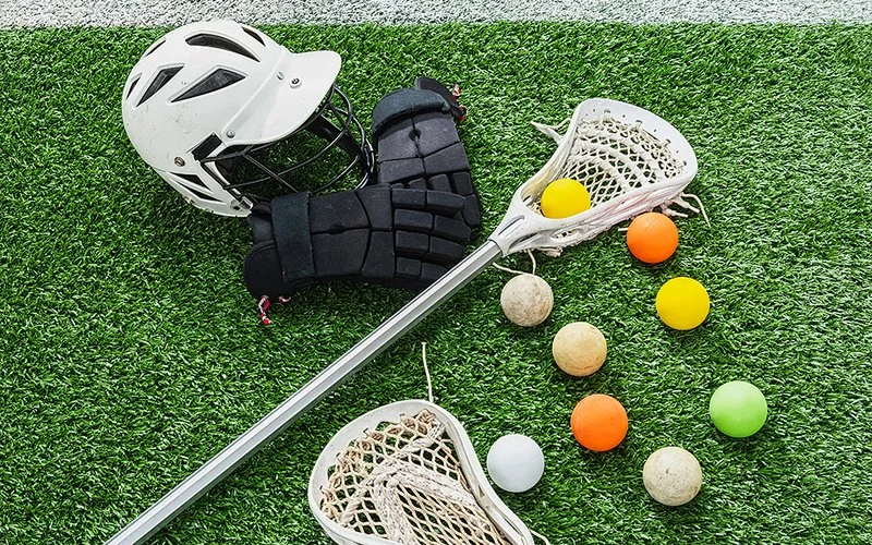 Auf einem Spielfeld liegt eine Lacrosse-Ausrüstung, bestehend aus Stöcken, Kugeln, Helm und Handschuhen.