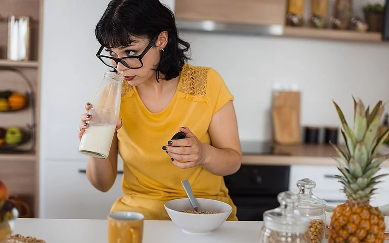 Eine jüngere Frau mit schwarzen mittellangen Haaren, schwarzer Brille und gelbem T-Shirt steht in einer Küche vor einer Schüssel Müsli und riecht an Milch in einer Flasche.