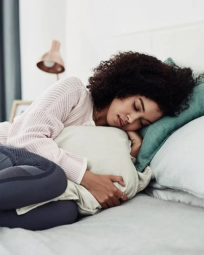 Eine Frau liegt mit angezogenen Beinen und einem Kissen, das sie sich vor den Bauch hält, auf einem Bett.