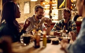 Ein lachender Mann mit einem Burger in der Hand sitzt abends mit Freunden an einem Tisch in einem Restaurant.