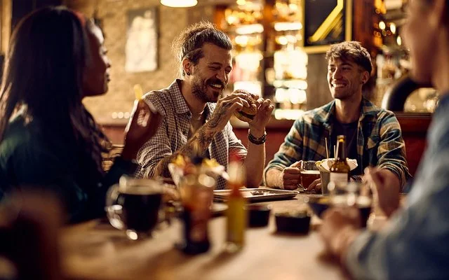 Ein lachender Mann mit einem Burger in der Hand sitzt abends mit Freunden an einem Tisch in einem Restaurant.