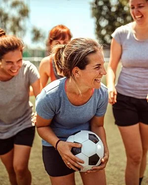 Eine Gruppe von Frauen in Sportkleidung steht lachend auf einem Fußballfeld. Die Frau in der Mitte hält einen Fußball. 