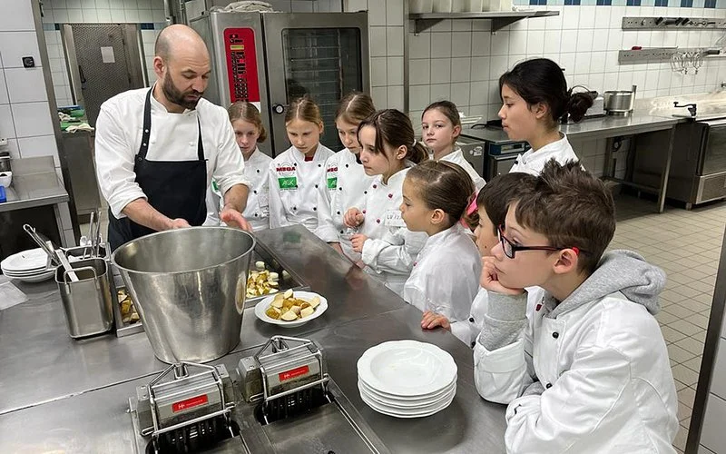 Mehrere Kinder stehen an der Kochinsel einer Gastro-Küche. Ein Koch erklärt die Zubereitung eines Gerichts.  
