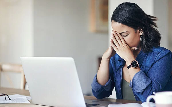 Eine junge Frau sitzt am Tisch vor einem Laptop und stützt das Gesicht in die Hände, die Augen geschlossen.