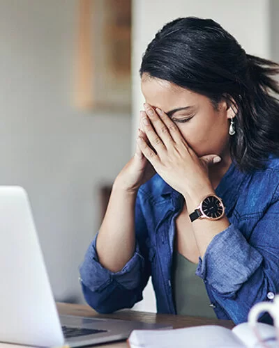 Eine junge Frau sitzt am Tisch vor einem Laptop und stützt das Gesicht in die Hände, die Augen geschlossen.