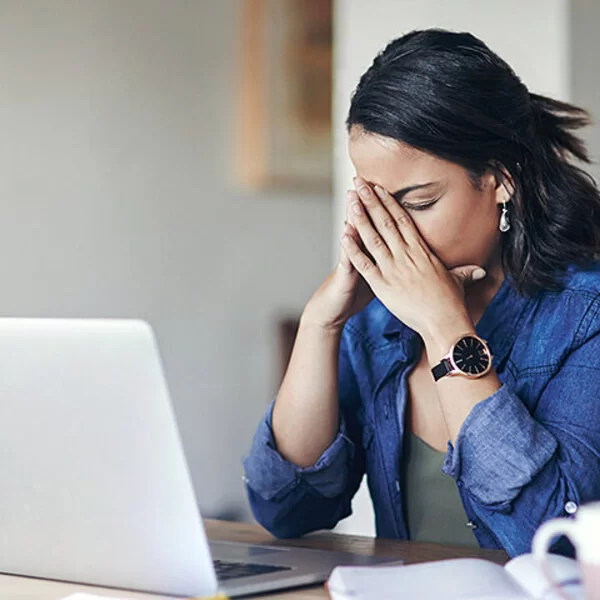 Eine junge Frau sitzt am Tisch vor einem Laptop und stützt das Gesicht in die Hände, die Augen geschlossen.