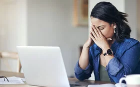Eine junge Frau sitzt am Tisch vor einem Laptop und stützt das Gesicht in die Hände, die Augen geschlossen.