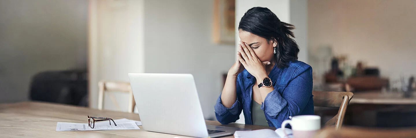 Eine junge Frau sitzt am Tisch vor einem Laptop und stützt das Gesicht in die Hände, die Augen geschlossen.