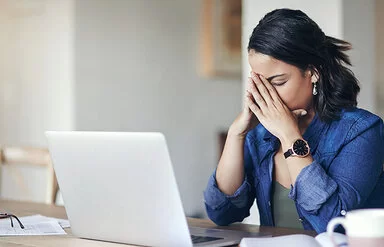 Eine junge Frau sitzt am Tisch vor einem Laptop und stützt das Gesicht in die Hände, die Augen geschlossen.