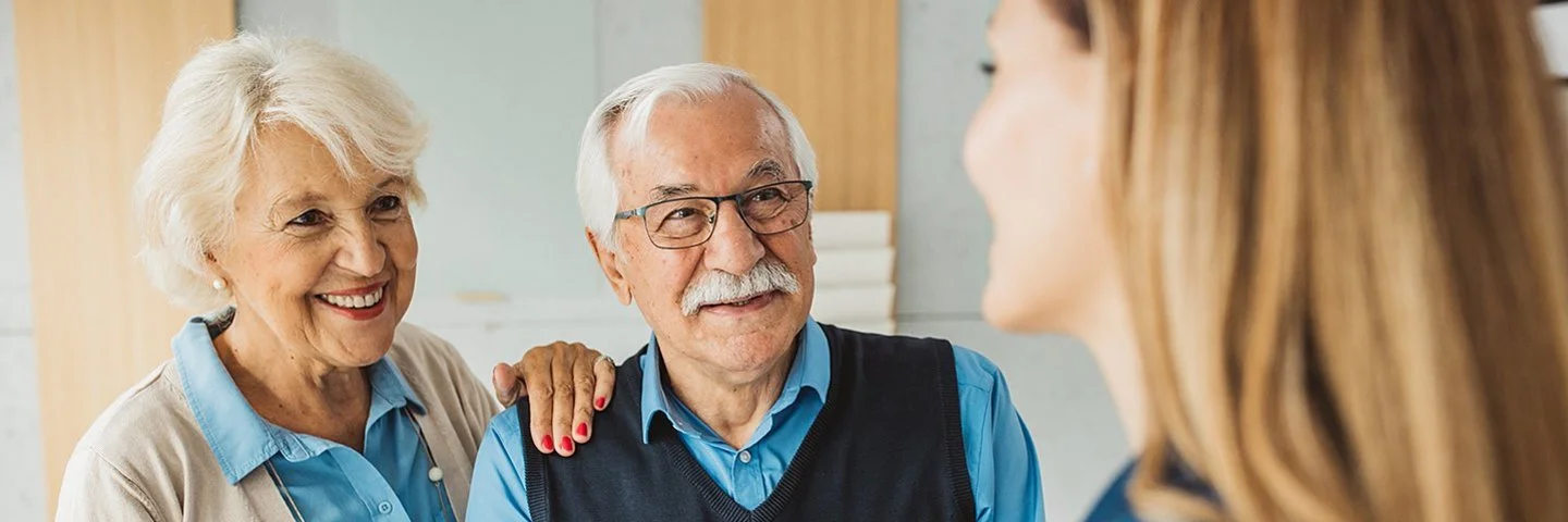Ein älteres Ehepaar steht in einem gemütlichen Eingangsbereich einer Wohnung. Sie empfangen eine Pflegerin, der der Mann freundlich die Hand reicht.