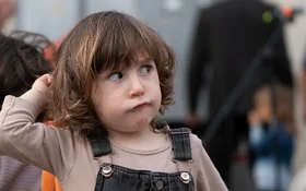 Ein Kleinkind mit braunen Haaren in einem Jeansoverall schaut zur Seite.