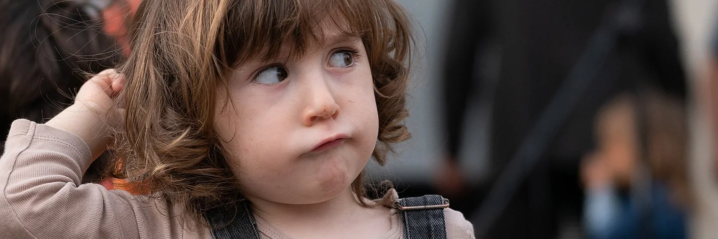 Ein Kleinkind mit braunen Haaren in einem Jeansoverall schaut zur Seite.