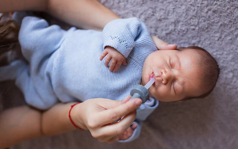 Eine Frau steckt eine Pipette in den Mund eines kleinen Jungen, der im blauen Strampler auf dem Rücken liegt.