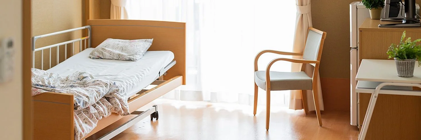 Pflegezimmer mit einem Pflegebett. Gardinen, Vorhänge, Topfpflanzen und ein Fernseher sorgen für eine gemütliche Atmosphäre.