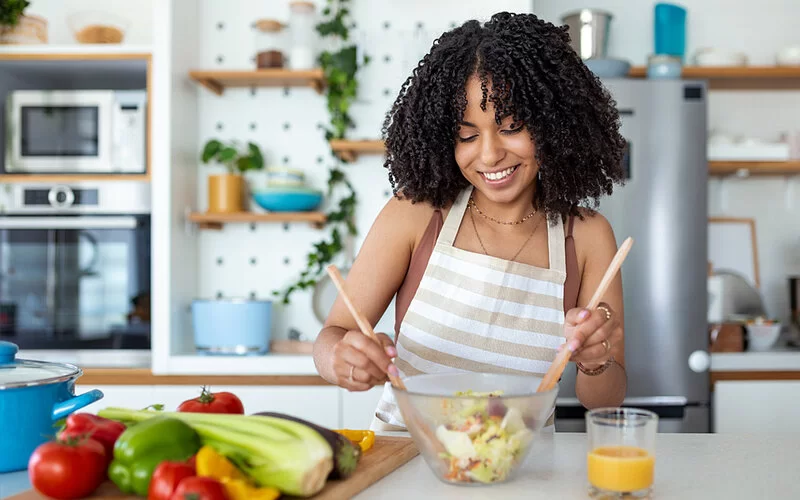 Eine junge Frau steht lächelnd in der Küche und richtet sich einen gemischten Salat an.