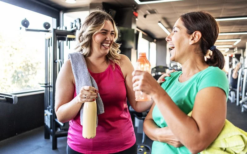 In einem Fitnessstudio unterhalten sich zwei Frauen in Sportkleidung lachend miteinander. Die eine fasst der anderen auf die Schulter. Sie halten jeweils eine Trinkflasche in der Hand und haben ein Handtuch dabei.