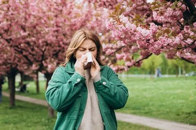 Eine junge Frau läuft durch einen grünen Park, in dem rosa Kirschblütenbäume blühen, und niest in ein Taschentuch.
