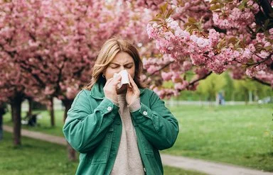 Eine junge Frau läuft durch einen grünen Park, in dem rosa Kirschblütenbäume blühen, und niest in ein Taschentuch.
