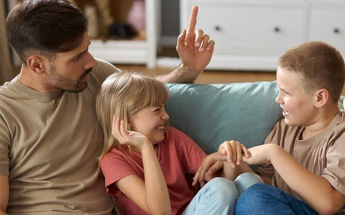 Ein Mann und eine Frau mit zwei Kindern im Teenageralter sitzen zusammen auf dem Sofa. Die Kinder albern übermütig herum, die Erwachsenen schauen ernst und angespannt.