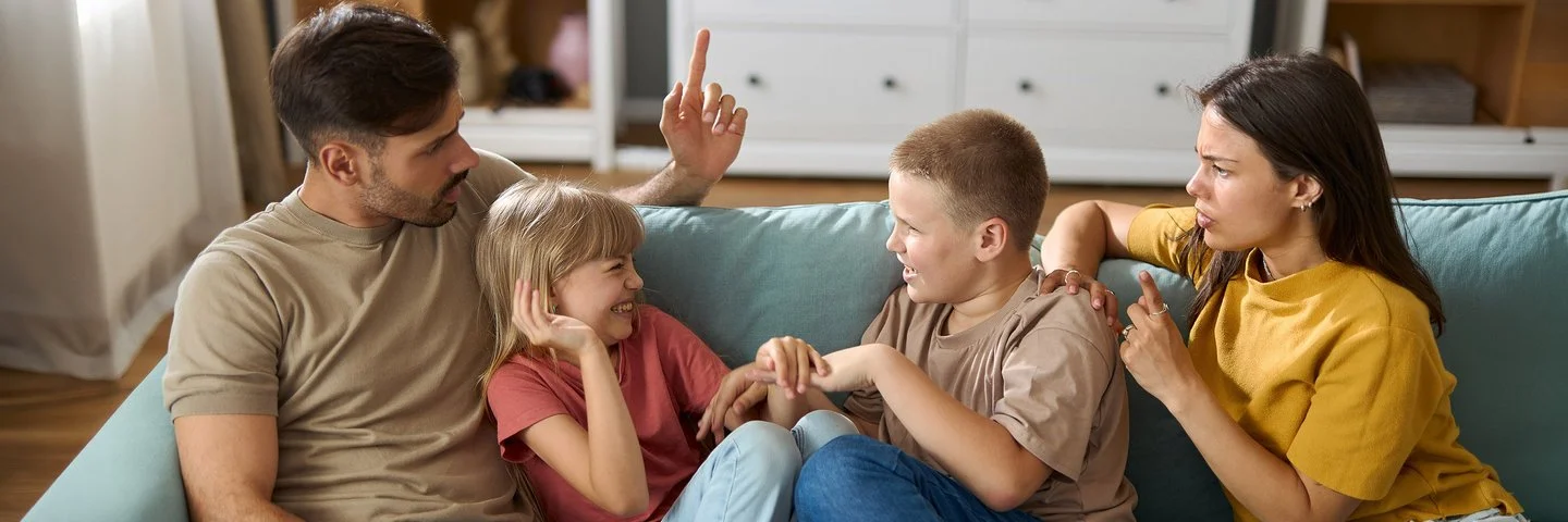 Ein Mann und eine Frau mit zwei Kindern im Teenageralter sitzen zusammen auf dem Sofa. Die Kinder albern übermütig herum, die Erwachsenen schauen ernst und angespannt.
