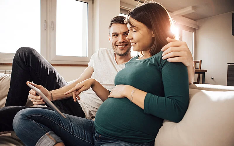 Schwangere junge Frau und ein junger Mann sitzen auf einem Sofa, er hat einen Arm um sie gelegt und beide schauen auf ein Tablet.