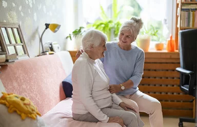 Eine Seniorin mit kurzen weißen Haaren, weißem Rollkragenpullover und grauer Hose sitzt auf dem Sofa. Neben ihr sitzt ihre Angehörige in blauem Langarmshirt und weißer Hose und stützt sie an Arm und Rücken.