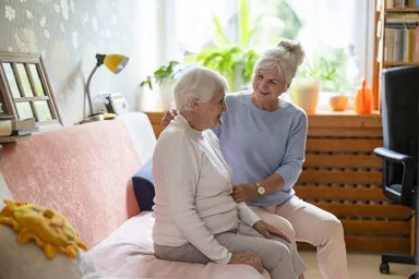 Eine Seniorin mit kurzen weißen Haaren, weißem Rollkragenpullover und grauer Hose sitzt auf dem Sofa. Neben ihr sitzt ihre Angehörige in blauem Langarmshirt und weißer Hose und stützt sie an Arm und Rücken.