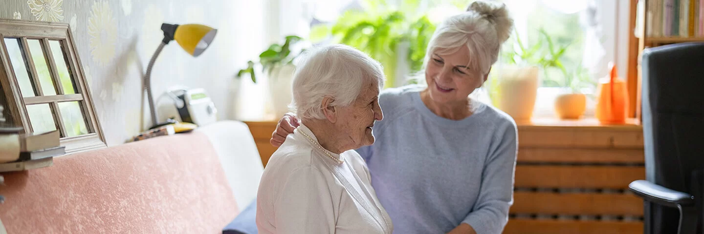 Eine Seniorin mit kurzen weißen Haaren, weißem Rollkragenpullover und grauer Hose sitzt auf dem Sofa. Neben ihr sitzt ihre Angehörige in blauem Langarmshirt und weißer Hose und stützt sie an Arm und Rücken.