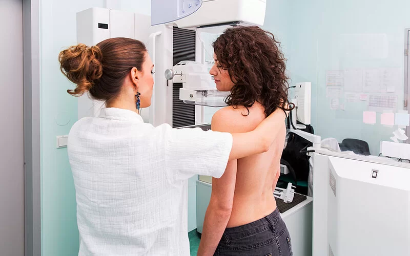 Eine Frau mit freiem Oberkörper steht an einem Mammographiegerät. Eine medizinische Fachkraft gibt Anweisungen.