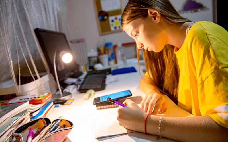 Ein Mädchen macht Hausaufgaben am Schreibtisch und lässt sich durch ihr Smartphone ablenken.