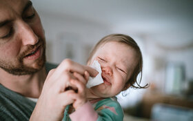 Ein Vater hält sein weinendes Kind mit Pseudokrupp auf dem Arm und wischt ihm mit einem Tuch den Mund ab.