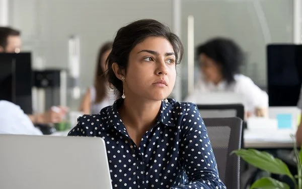 Eine jüngere Frau in blau-weiß gepunkteter Bluse sitzt im Großraumbüro vor ihrem Laptop und schaut nachdenklich.