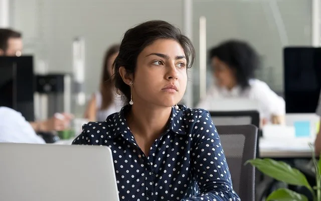 Eine jüngere Frau in blau-weiß gepunkteter Bluse sitzt im Großraumbüro vor ihrem Laptop und schaut nachdenklich.