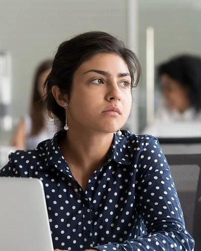 Eine jüngere Frau in blau-weiß gepunkteter Bluse sitzt im Großraumbüro vor ihrem Laptop und schaut nachdenklich.