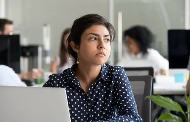 Eine jüngere Frau in blau-weiß gepunkteter Bluse sitzt im Großraumbüro vor ihrem Laptop und schaut nachdenklich.