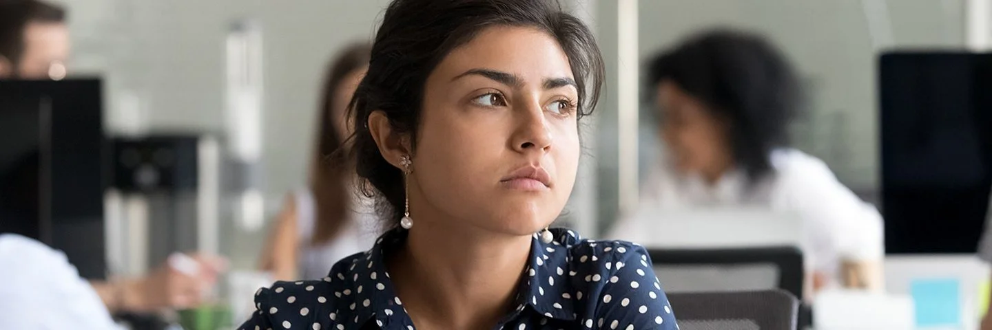 Eine jüngere Frau in blau-weiß gepunkteter Bluse sitzt im Großraumbüro vor ihrem Laptop und schaut nachdenklich.