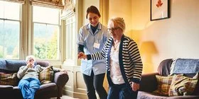 Eine Pflegefachfrau sützt eine ältere Dame an der rechten Körperseite ab, während diese ein paar Schritte geht.
