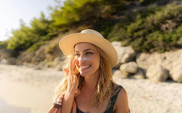 Eine lachende junge Frau mit Sonnenhut trägt am Strand Sonnencreme im Gesicht auf.