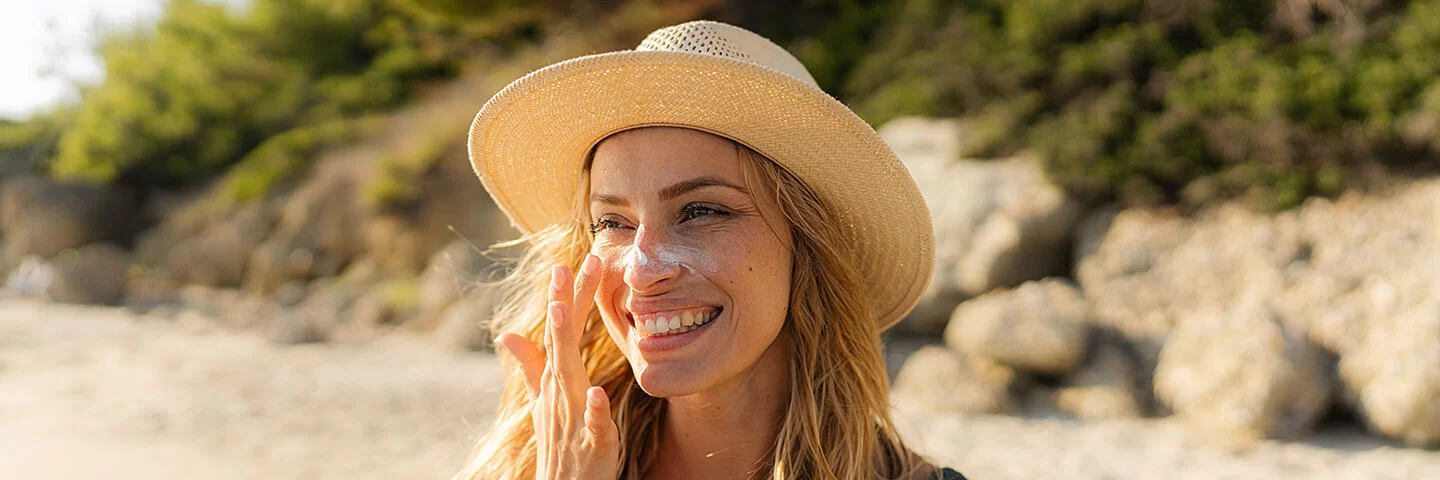 Eine lachende junge Frau mit Sonnenhut trägt am Strand Sonnencreme im Gesicht auf.