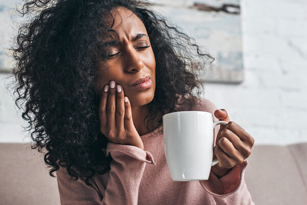 Eine Frau hat Zahnschmerzen nach einer Weisheitszahn-OP und hält eine Tasse mit Tee in der Hand.