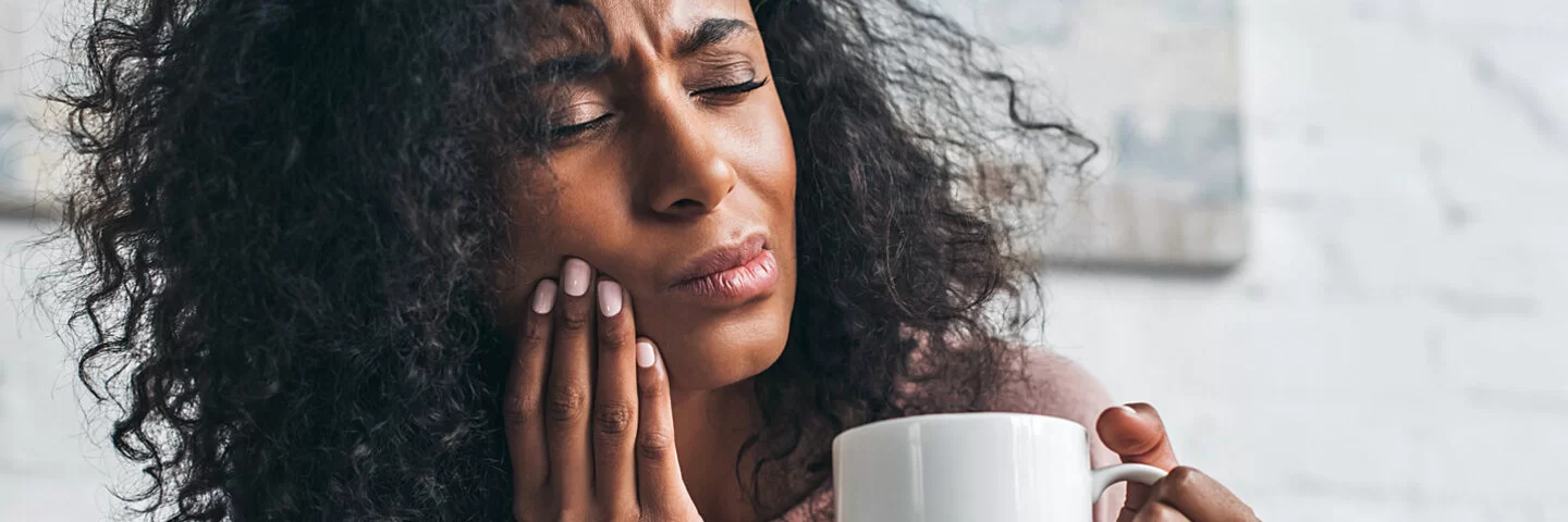 Eine Frau hat Zahnschmerzen nach einer Weisheitszahn-OP und hält eine Tasse mit Tee in der Hand.