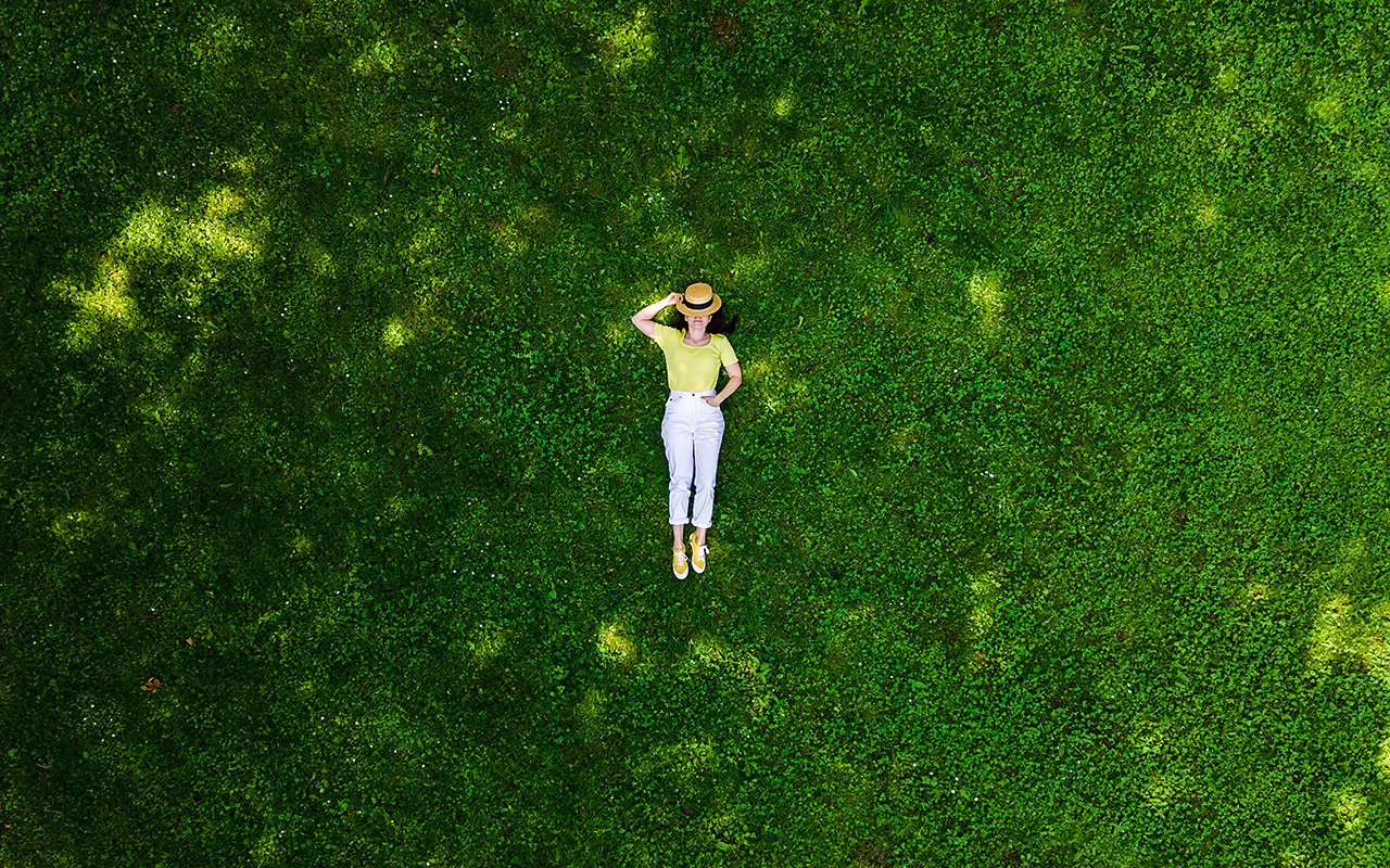 Person mit Strohhut und heller Kleidung liegt auf einer grünen Wiese und blickt nach oben.