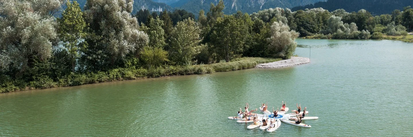 Personen sind mit ihren SUP Brettern auf einem See im Kreis und machen Yogaübungen. Im Hintergrund sind die Berge.