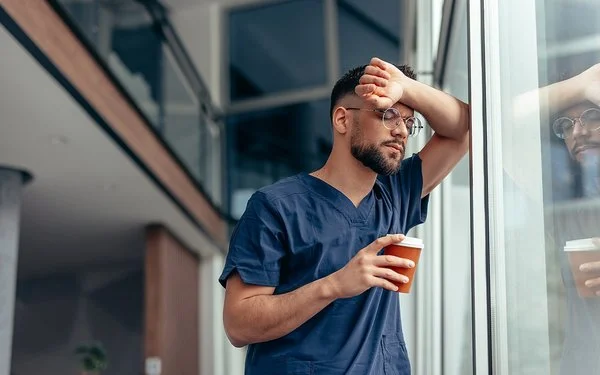 Ein junger Mann in medizinischer Arbeitskleidung lehnt mit einem Ellenbogen an einem Fenster, und stützt dabei die Stirn auf den erhobenen Unterarm. In der anderen Hand hält er einen Becher. Seine Körperhaltung wirkt erschöpft und nachdenklich.