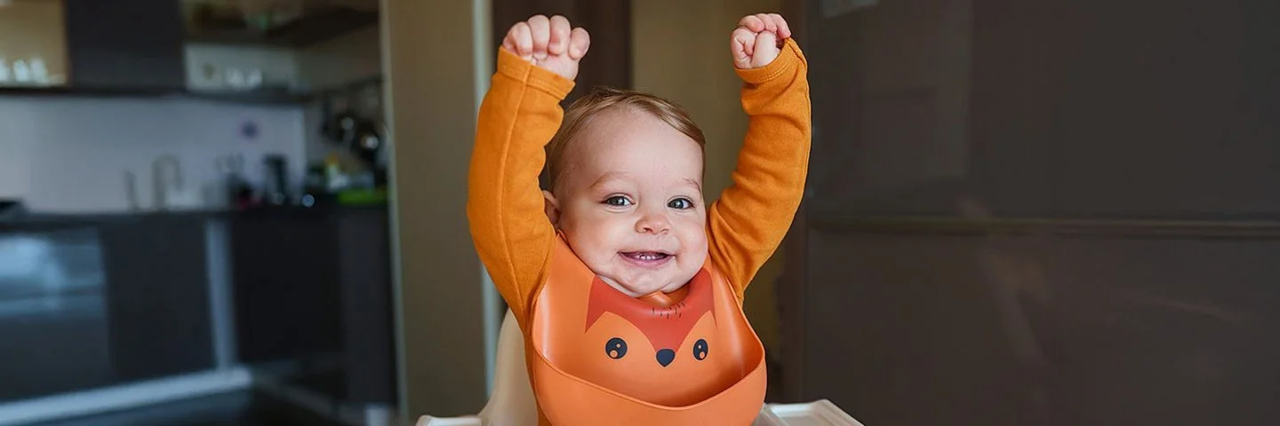 Ein Baby in einem orangefarbenen Pulli sitzt in einem Hochstuhl, lacht in die Kamera und hält die Arme nach oben über seinen Kopf.