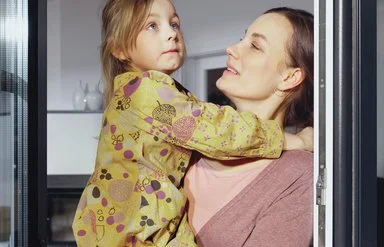 Eine Frau hält Kind auf dem Arm vor einem Fenster, beide blicken nach oben
